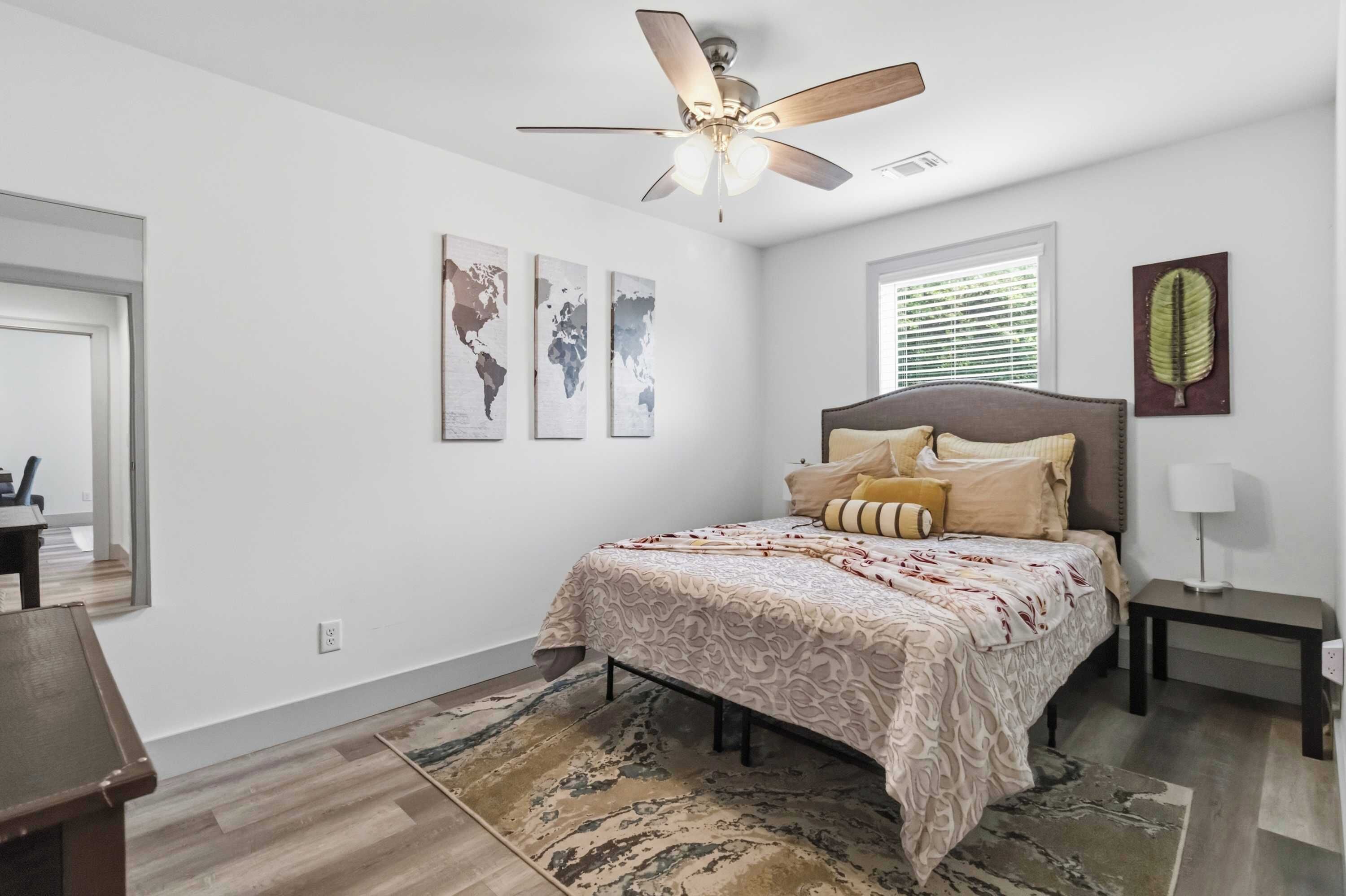 A bedroom with a bed, a desk, a chair, a ceiling fan, a window with blinds, and three wall-mounted world maps.