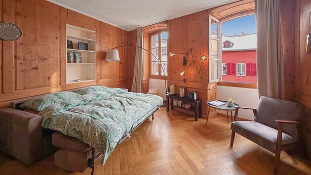 A cozy room with a bed, a chair, a small table, and a window with a view of buildings. - The One and Only in Sion (Sion)