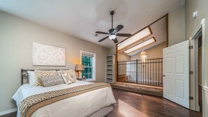 A bedroom with a large bed, a ceiling fan, a window, and a staircase leading to an upper level.