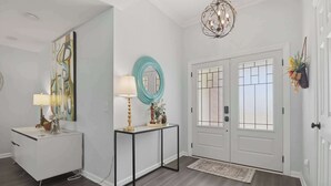 A hallway with a white console table, a round mirror, a black console table, a white door with a glass panel, and a chandelier. - Niagara Retreat in the Heart of NWA (Springdale)