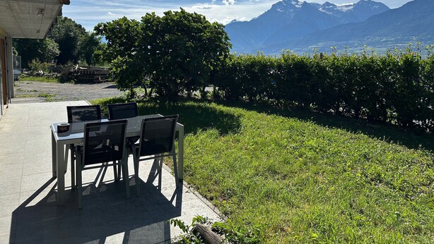 An outdoor seating area with a table and chairs, a building with a balcony, and a mountain view.