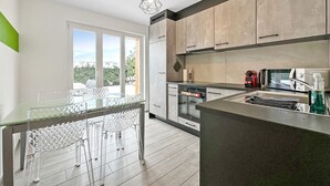 A modern kitchen with a dining area, large windows, and a view of greenery.