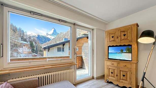 Ein Zimmer mit einem großen Fenster, einem Holfachschrank mit Fernseher, einem Heizkörper und einer Bank.