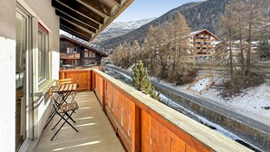 Ein Holzbalkon mit Tisch und Stühlen, der einen schneebedeckten Talblick und ein Gebäude mit mehreren Balkonen bietet.