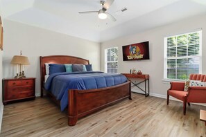 A bedroom with a wooden bed, a nightstand, a chair, a desk, a ceiling fan, and a window with blinds.