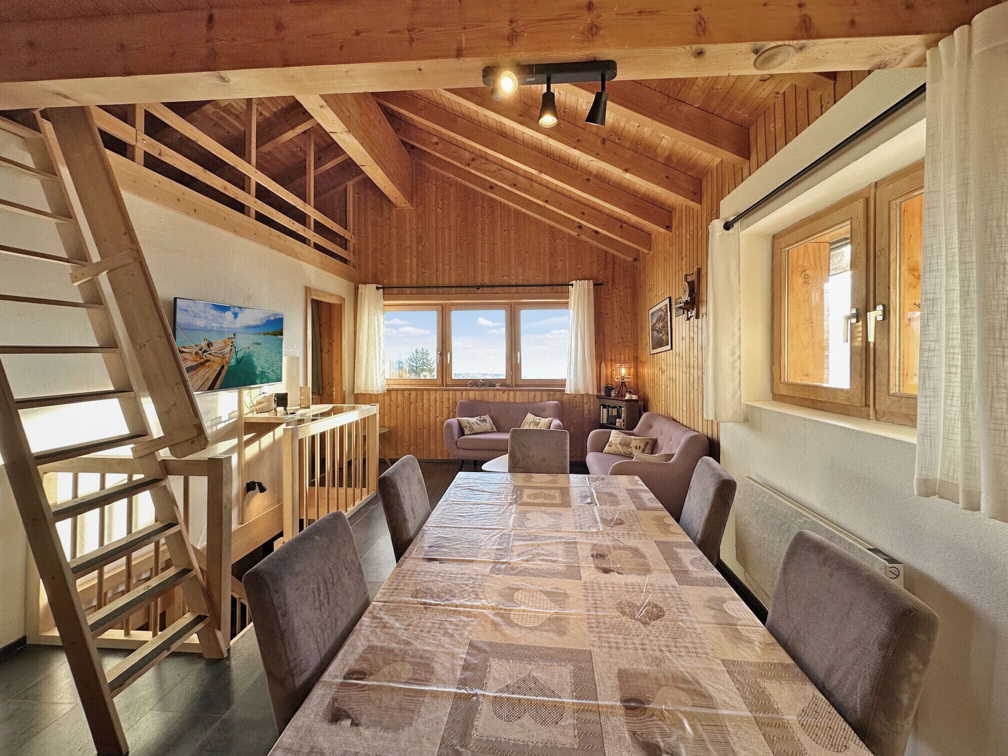 A wooden interior with a dining table, chairs, a sofa, and a TV mounted on the wall.