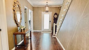 A hallway with wooden flooring, a mirror, a console table with a vase, a chandelier, and a clock. - Enjoy 3 King beds with ensuite baths, 8 total beds (Fayetteville)