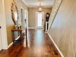 A hallway with wooden flooring, a mirror, a console table with a vase, a chandelier, and a clock. - Enjoy 3 King beds with ensuite baths, 8 total beds (Fayetteville)