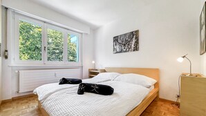 Un dormitorio con un ventanal grande, una cama con ropa de cama blanca, una mesita de noche con una lámpara y un cuadro en la pared.