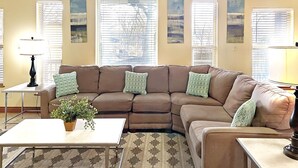 A living room with a brown sectional sofa, a white coffee table, and a patterned rug. There are two lamps on side tables, and a plant on the coffee table.