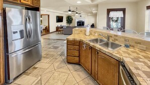 A modern kitchen with a stainless steel refrigerator, wooden cabinets, a large island with a sink, and a tiled floor.