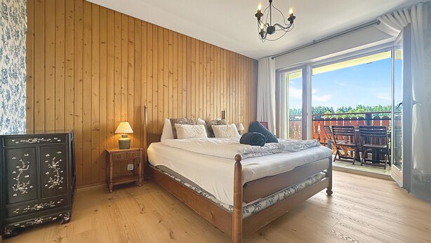 Ein Schlafzimmer mit Holzboden, einem Bett mit weißer Bettwäsche, einem Nachttisch mit Lampe und Blick ins Freie durch ein großes Fenster.