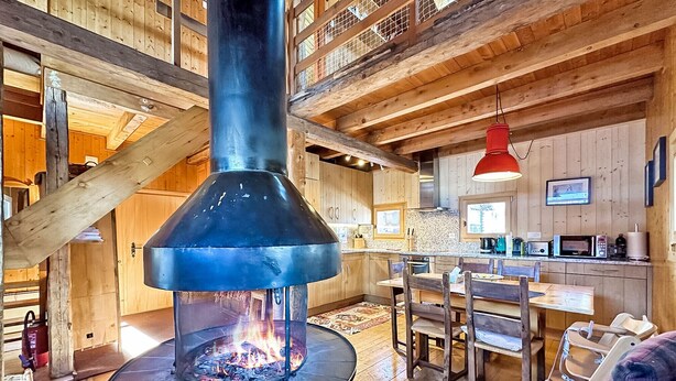 A cozy cabin interior with a central wood-burning stove, wooden beams, a dining area, and a kitchen with modern appliances.