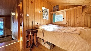 A wooden cabin room with a bed, a nightstand, a window with a view, and a wall-mounted lamp.