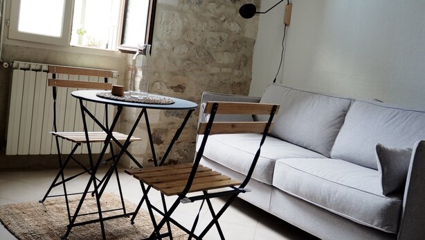 A room with a stone wall, a grey sofa, a wooden table with two chairs, and a radiator. - Quiet nest with terrace in the city center (Avignon)