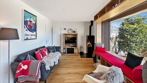 Ein modernes Wohnzimmer mit Kamin, einer Sofagarnitur, einem Fernseher und einem großen Fenster mit Blick auf Bäume.