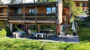 Una cabaña de madera de dos pisos con un patio, mobiliario exterior y vistas del bosque y las montañas circundantes.