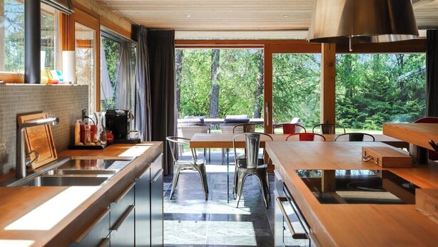 Una cocina moderna con un amplio ventanal, armarios de madera y una zona de comedor con sillas metálicas.