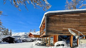 Una cabaña de madera con entrada de vidrio, rodeada de suelo y árboles cubiertos de nieve.