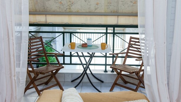 A balcony with two wooden chairs, a small table with two yellow cups, and a view of greenery. - Cozy apt in Peristeri near Metro station (Peristeri)