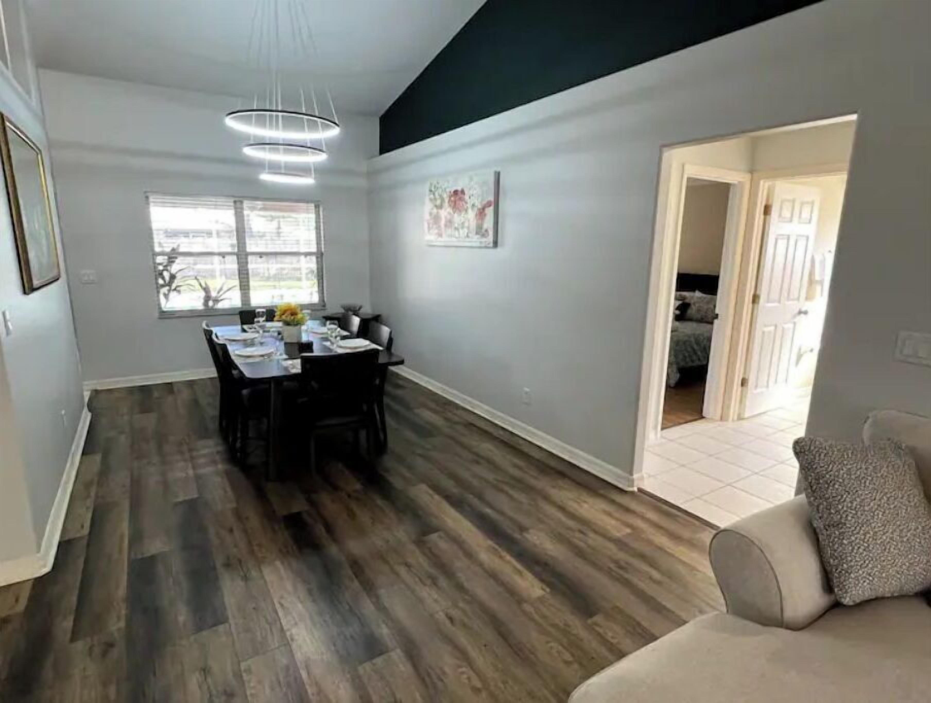 Un vaste espace de vie comprenant une table à manger pour quatre personnes, un canapé gris et une porte-fenêtre donnant sur une chambre.