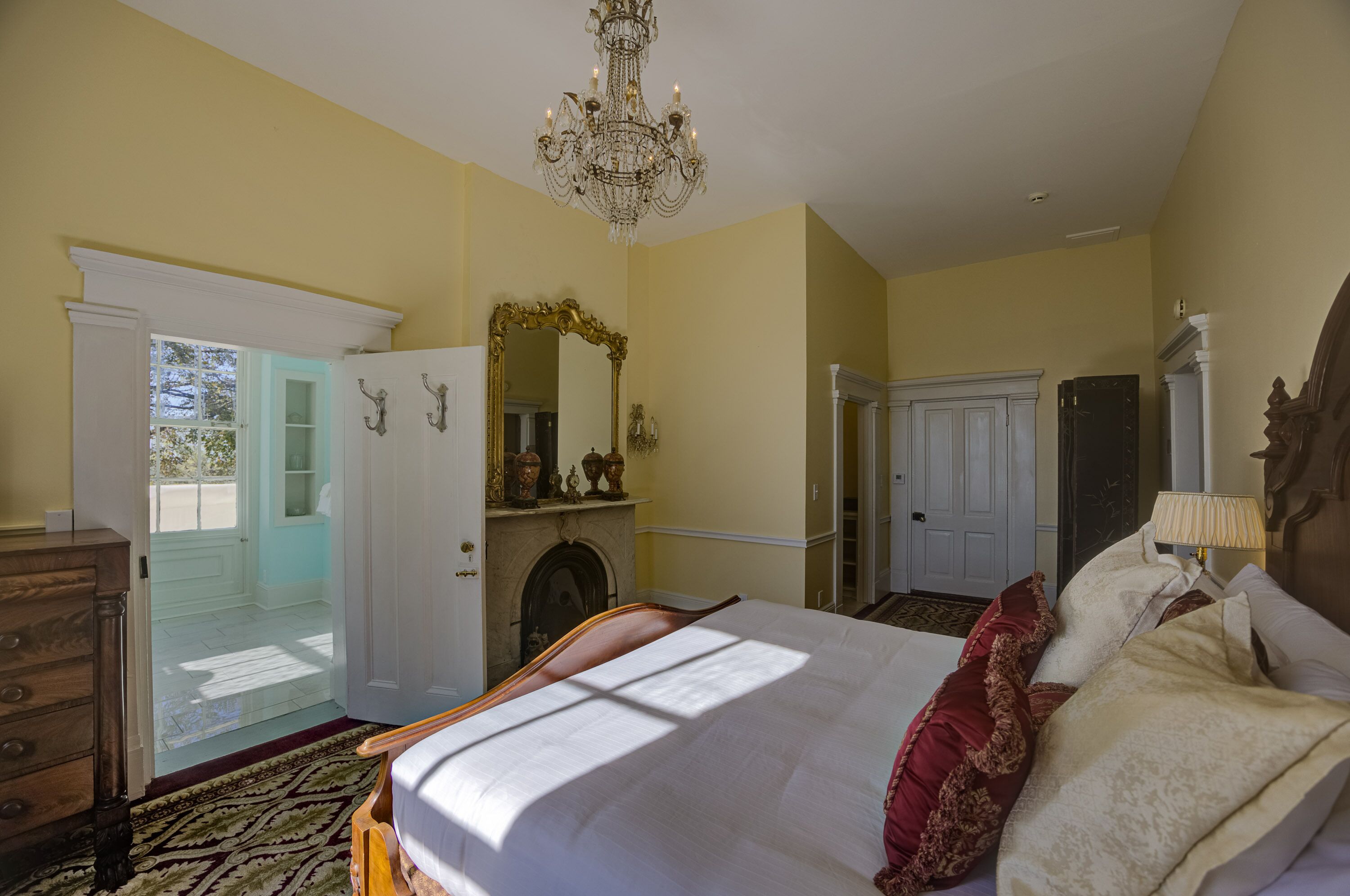 Une chambre à coucher comprenant un lit, une commode, un miroir, un lustre et une porte donnant sur un balcon.