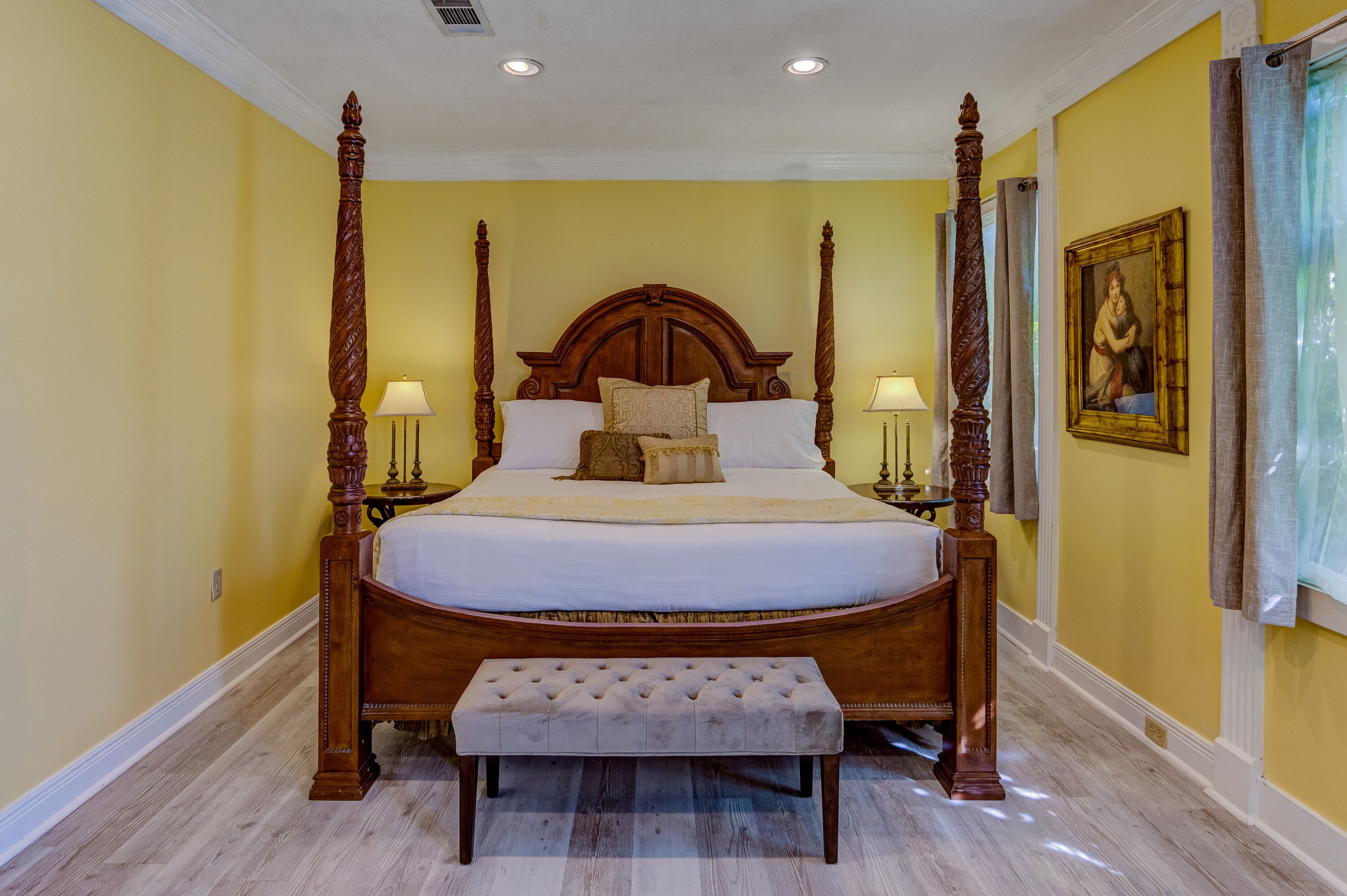 A four-poster bed with a tufted bench in front, a nightstand with a lamp, and a framed painting on the wall.
