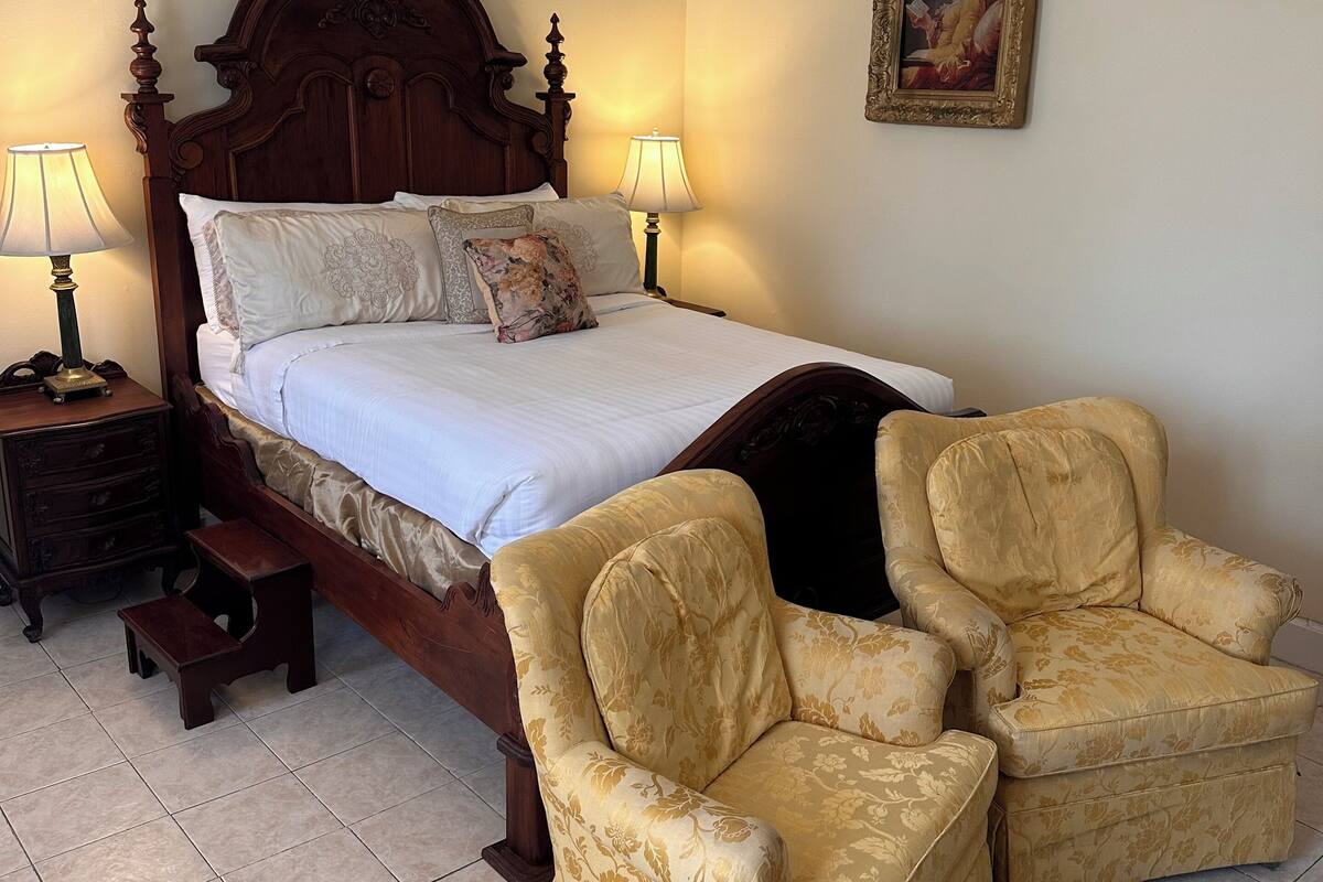 A bedroom with a wooden bed, two bedside lamps, a nightstand, a small wooden stool, and two upholstered chairs.