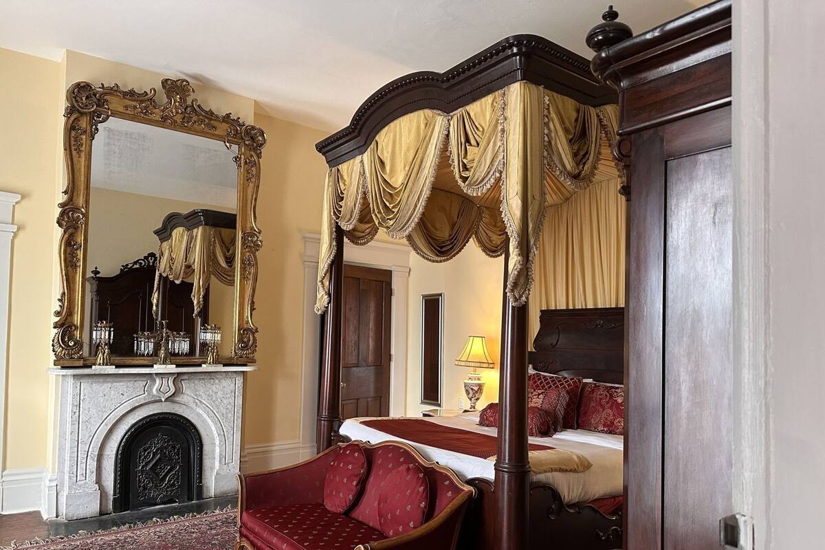 A four-poster canopy bed with a red upholstered chair, a large ornate mirror, and a fireplace.