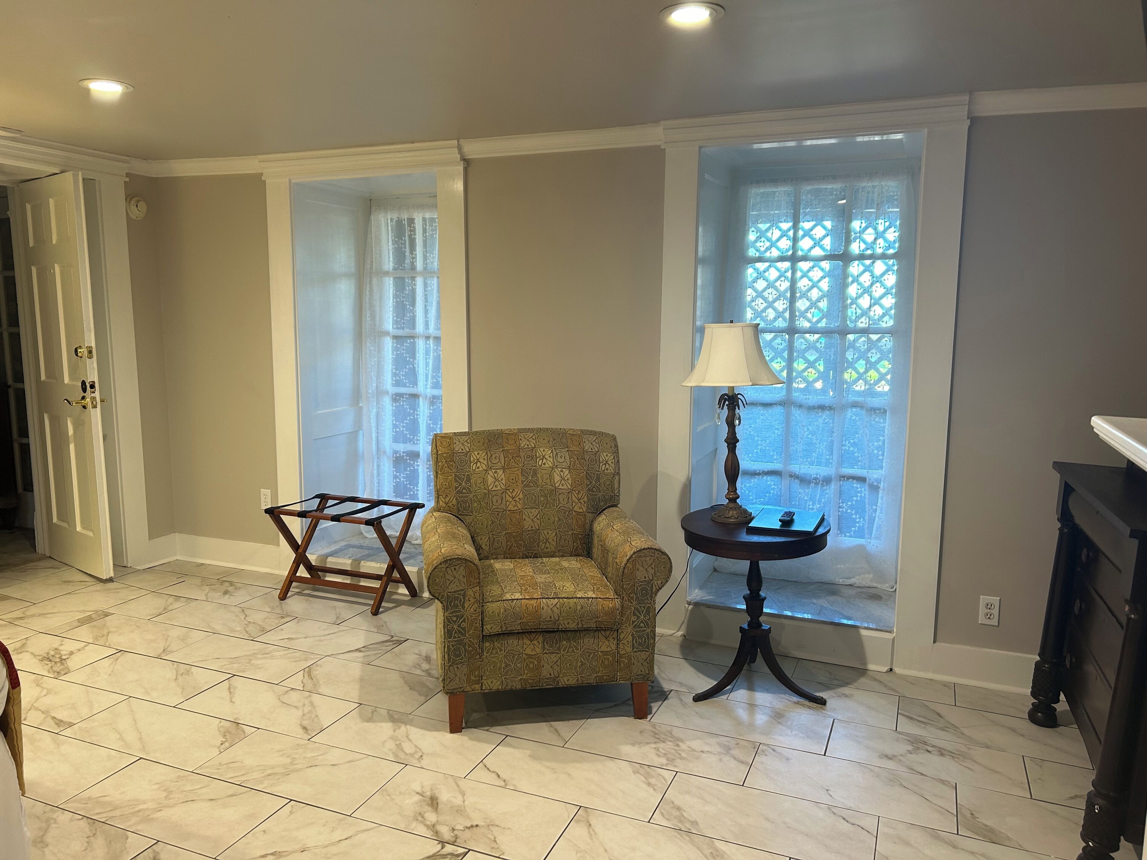 A room with a patterned armchair, a small table with a lamp, and a window with a lattice design.