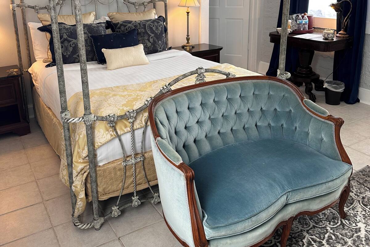 A four-poster bed with a tufted headboard, a tufted armchair, a nightstand with a lamp, and a desk with a lamp and a cup.