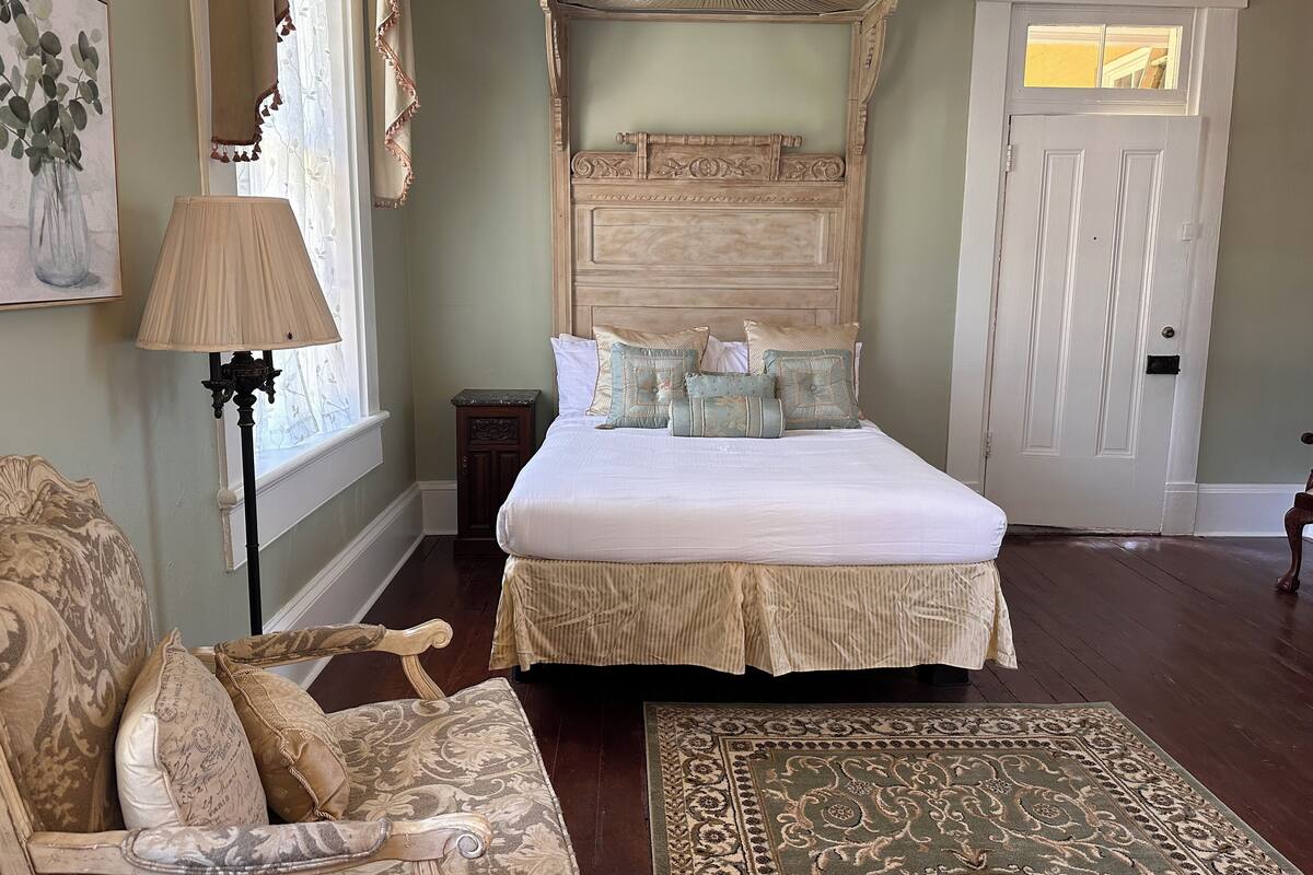 A bedroom with a large bed, a nightstand, a chair, a lamp, and a door.
