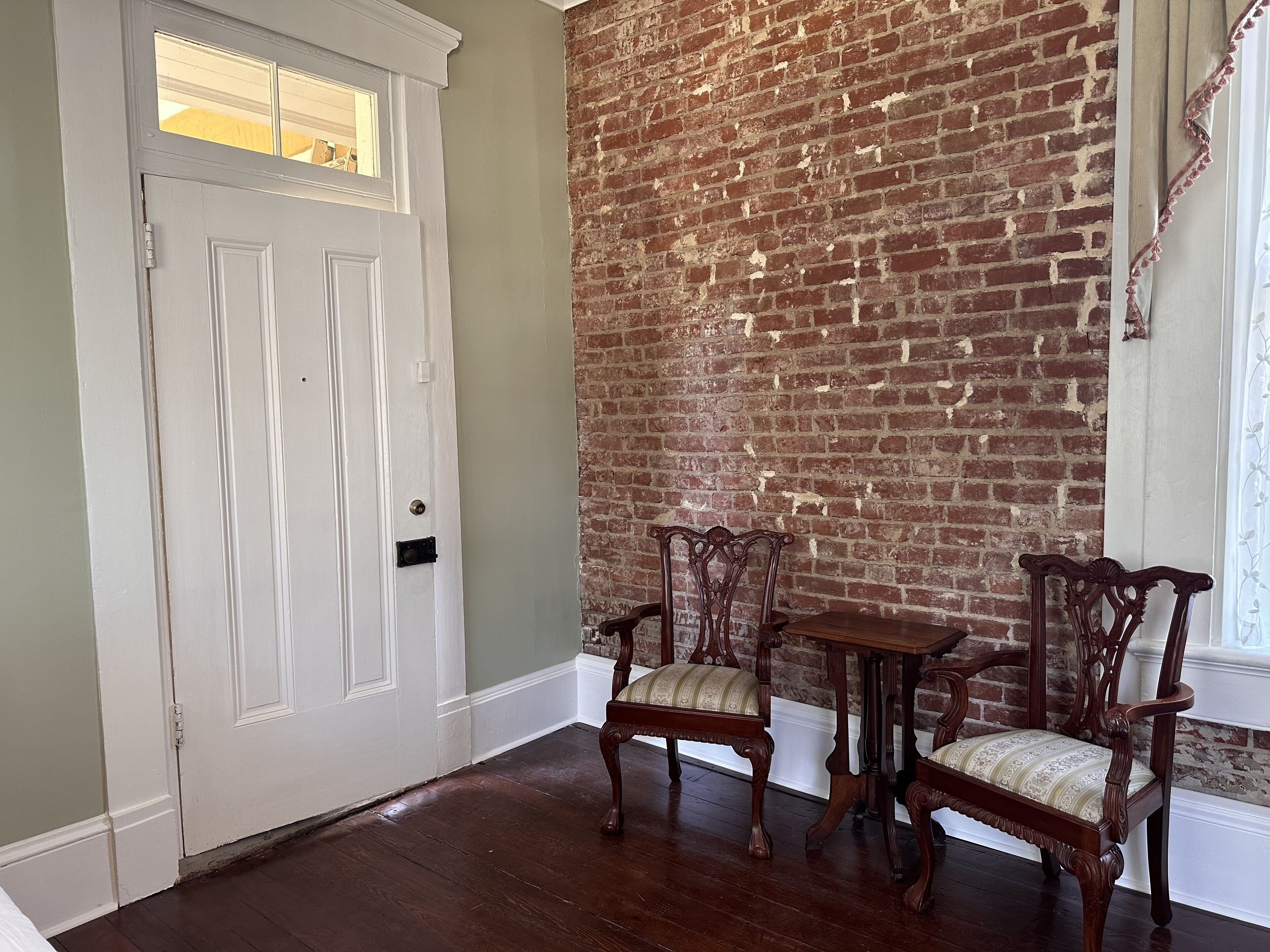 A room with a white door, a brick wall, and a wooden floor. There is a small table with two chairs.