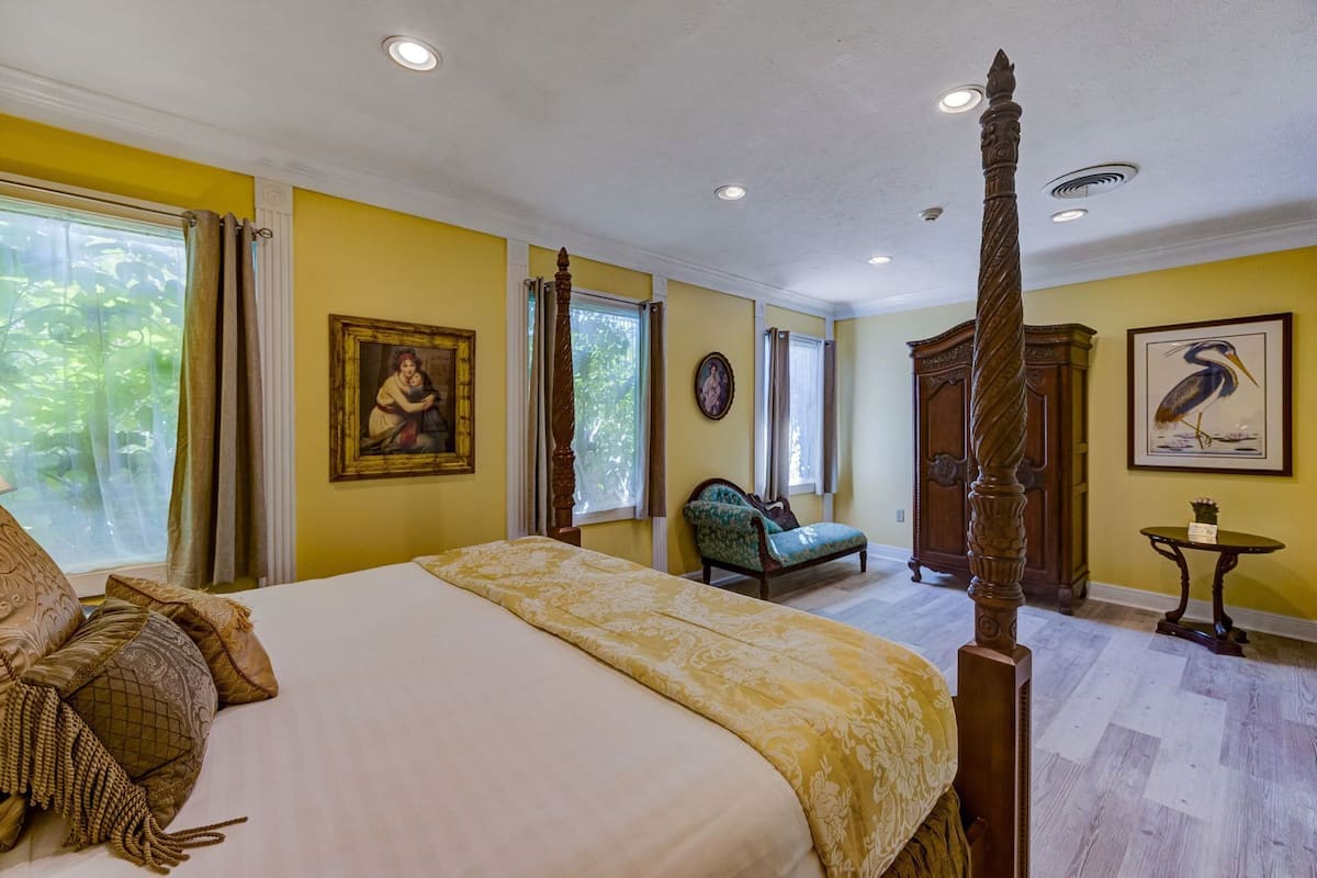 A bedroom with a four-poster bed, a chair, a wooden cabinet, and artwork on the walls.