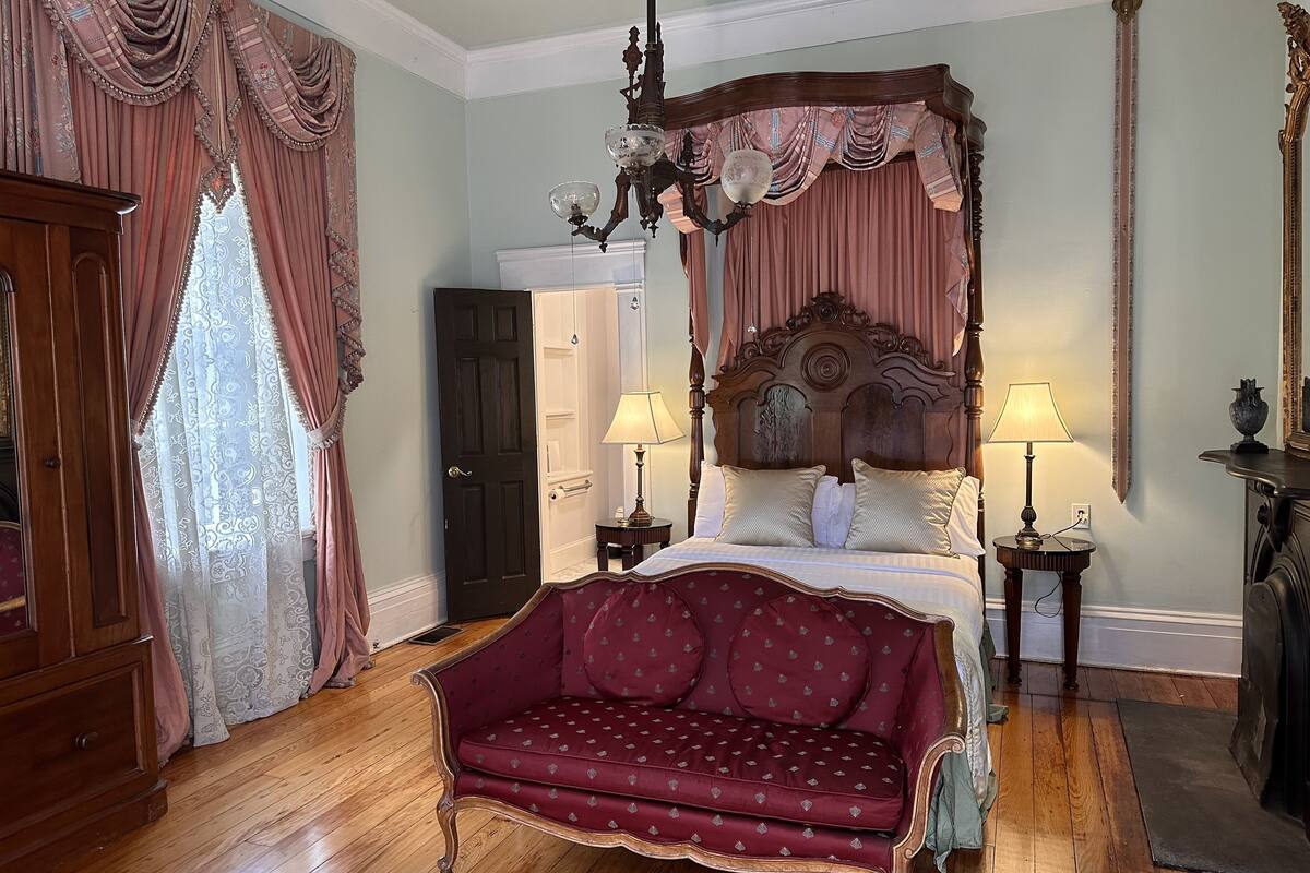 A classic four-poster bed with a wooden headboard, two bedside tables with lamps, a red upholstered sofa, and a wooden wardrobe.