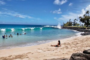 Ein Strand mit klarem, blauem Wasser, Menschen beim Schwimmen und Surfen, und eine Frau sitzt im Sand.