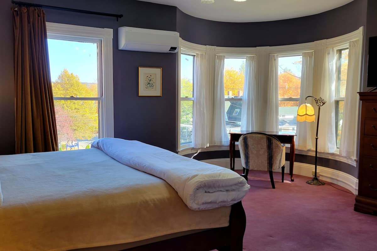 Une chambre à coucher avec un grand lit, un bureau, une chaise et une commode. Plusieurs fenêtres offrent une vue sur l’extérieur.