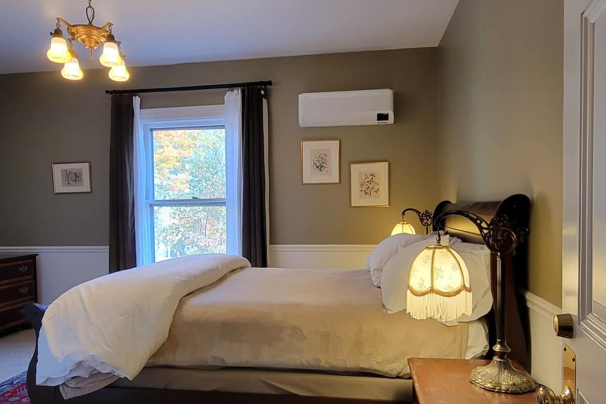 Une chambre à coucher avec un grand lit, une table de chevet avec une lampe, une fenêtre avec des rideaux et un luminaire au plafond.