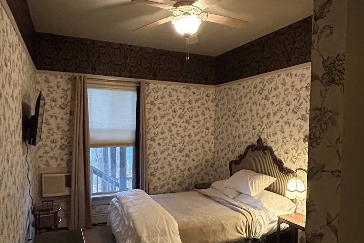 Une chambre à coucher comprenant un lit, un ventilateur de plafond, une table de chevet avec une lampe, une fenêtre avec des rideaux et une petite table.