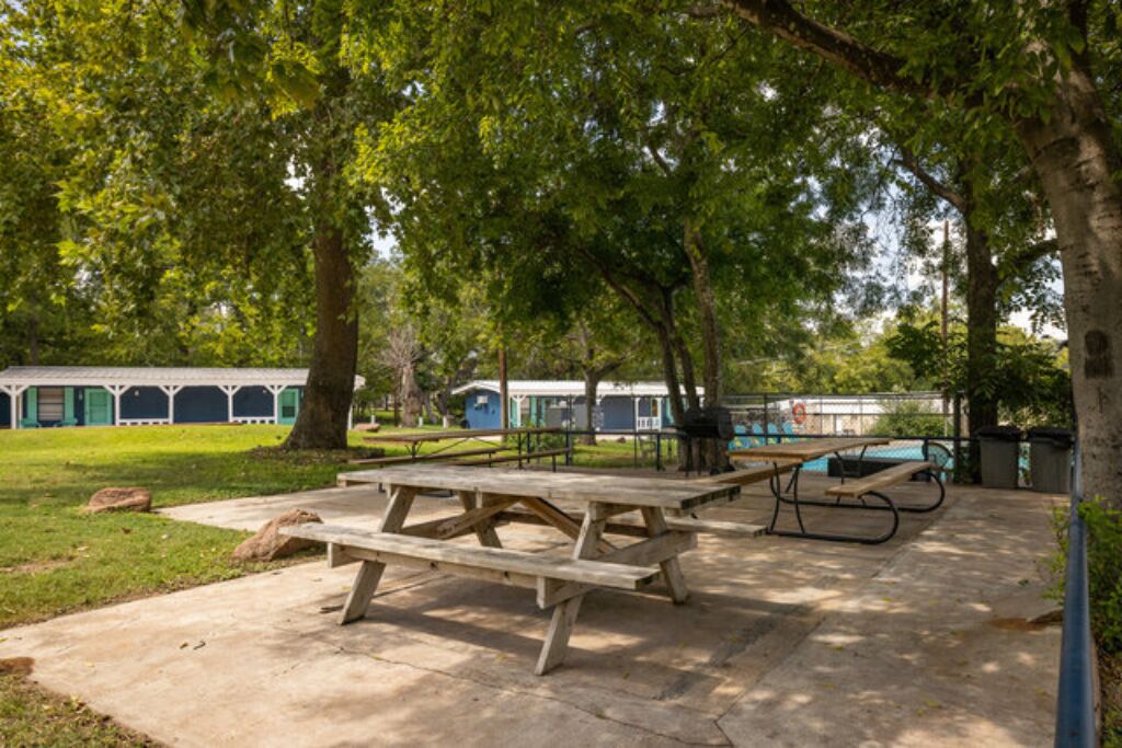 Un área de picnic al aire libre con mesas y una piscina, y un edificio con un paseo techado al fondo.