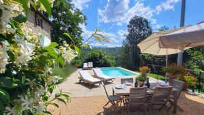 Zona de piscina exterior con sillones de descanso y una mesa lista para comer, rodeada de follaje y un edificio de piedra.