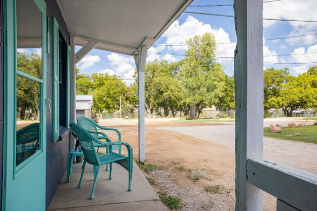 A covered porch with two green chairs, a concrete floor, and a view of trees and a dirt path.
