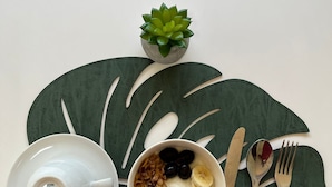 A breakfast setup with a bowl of cereal, bananas, and berries, a teapot, and cutlery on a leaf-shaped placemat.