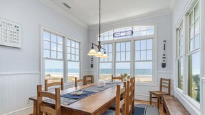 A dining area with a wooden table and chairs, large windows with ocean views, and a hanging light fixture. - Palmettos & Sunshine: Oceanfront w/ Pool, Hot Tub, & Stunning Views (Garden City Beach)