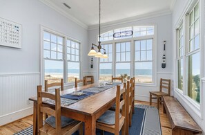 Un coin repas avec une table et des chaises en bois, de grandes fenêtres donnant sur l’océan et un luminaire suspendu.