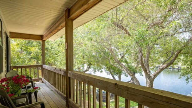 Une terrasse en bois avec des chaises et une table, donnant sur un lac bordé d’arbres et sur un quai.
