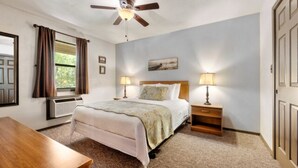Une chambre à coucher comprenant un lit, deux tables de chevet avec des lampes, un ventilateur de plafond, une fenêtre avec des rideaux et une porte.