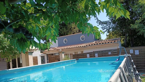 A swimming pool with clear blue water, surrounded by trees and a building with a tiled roof. - The house around the fig tree (Aramon)