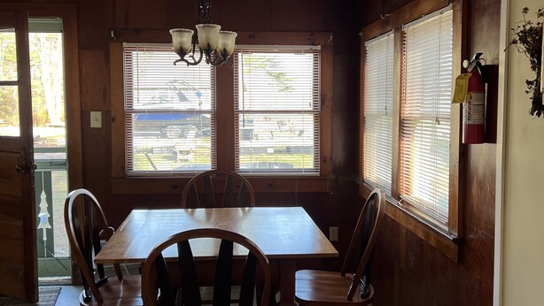A dining area with a wooden table and chairs, a chandelier, and windows with blinds.