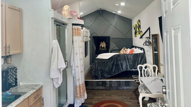 Un dormitorio pequeño con cama, una mesita, una silla, un espejo y zona de ducha.